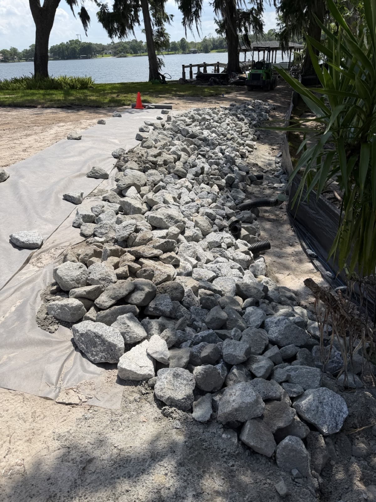 Graded granite riprap being placed over filter fabric along a Central Florida lakefront shoreline, with a stormwater outfall pipe and silt fence protecting the lake during installation